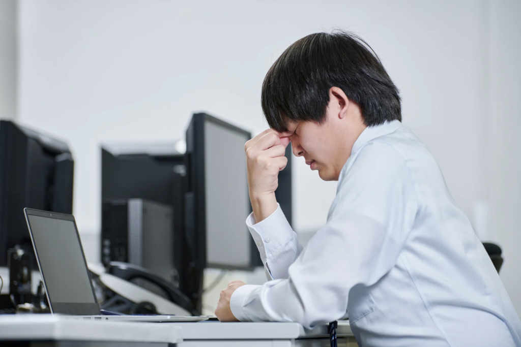 Conflict De-Escalation: A person in a white shirt sits at a desk with their head in their hand in front of computer screens.